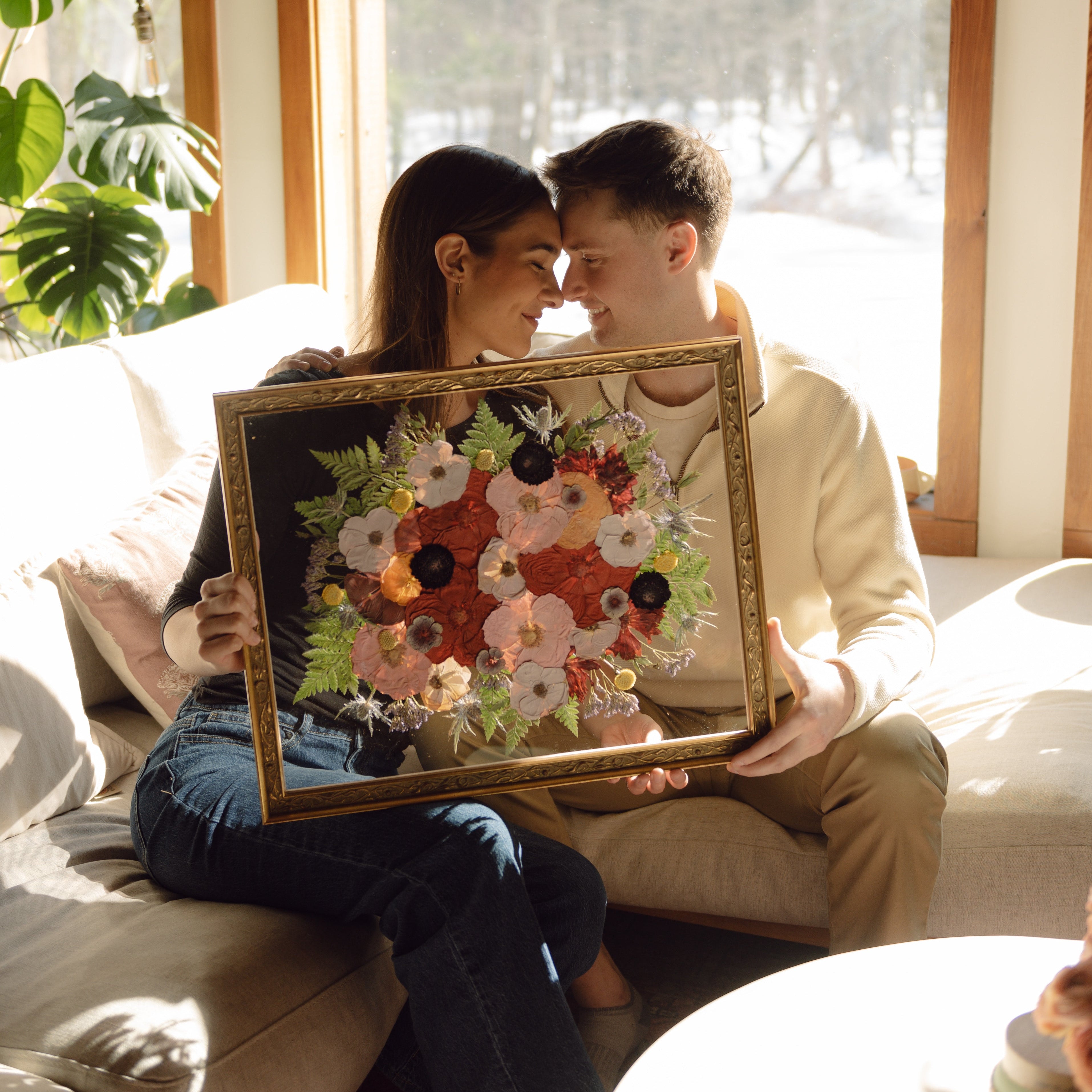 Two people sitting on a couch holding a framed floral artwork in a bright room with large windows.