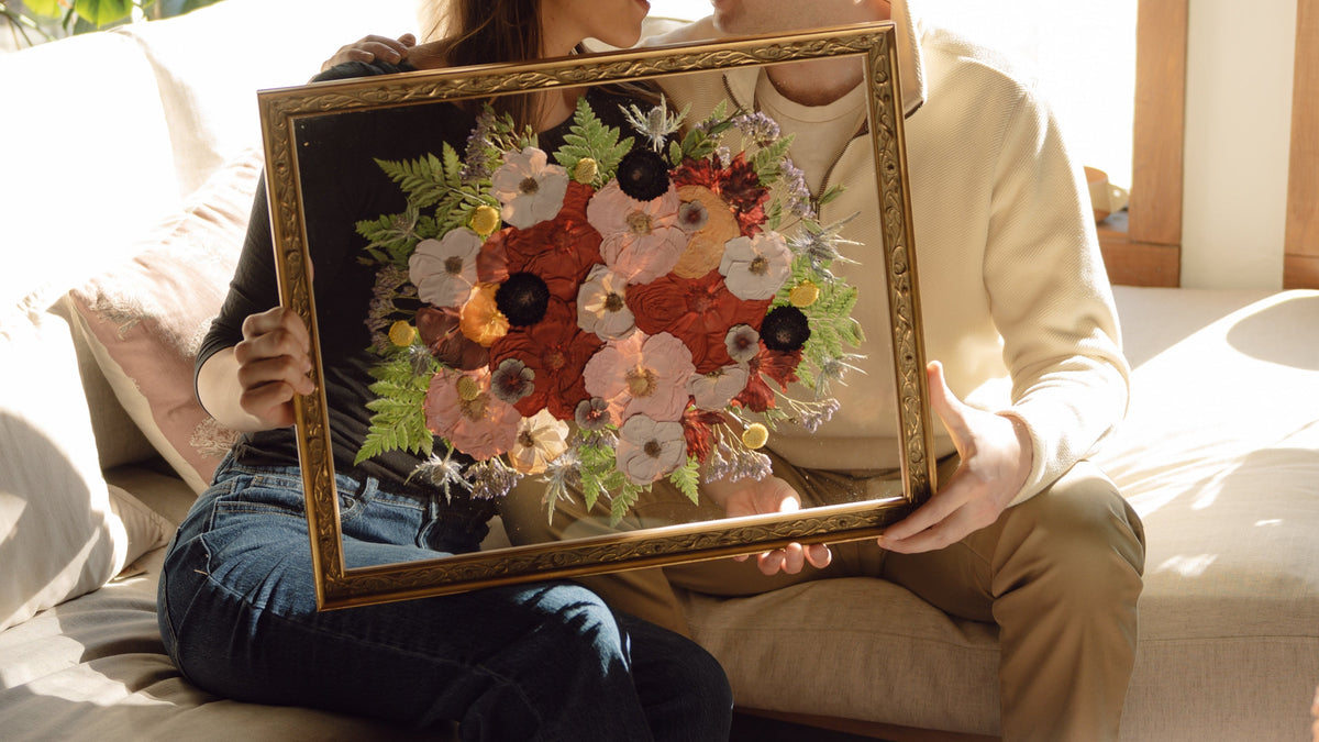 Two people sitting on a couch holding a framed floral artwork in a bright room with large windows.
