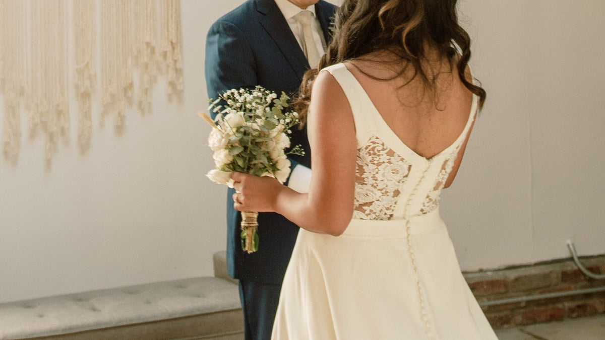 A bride in a white lace wedding gown with a backless V-neck and a groom in a navy suit standing in a bright, modern indoor space with a macrame wall hanging, bench, and patterned rug.