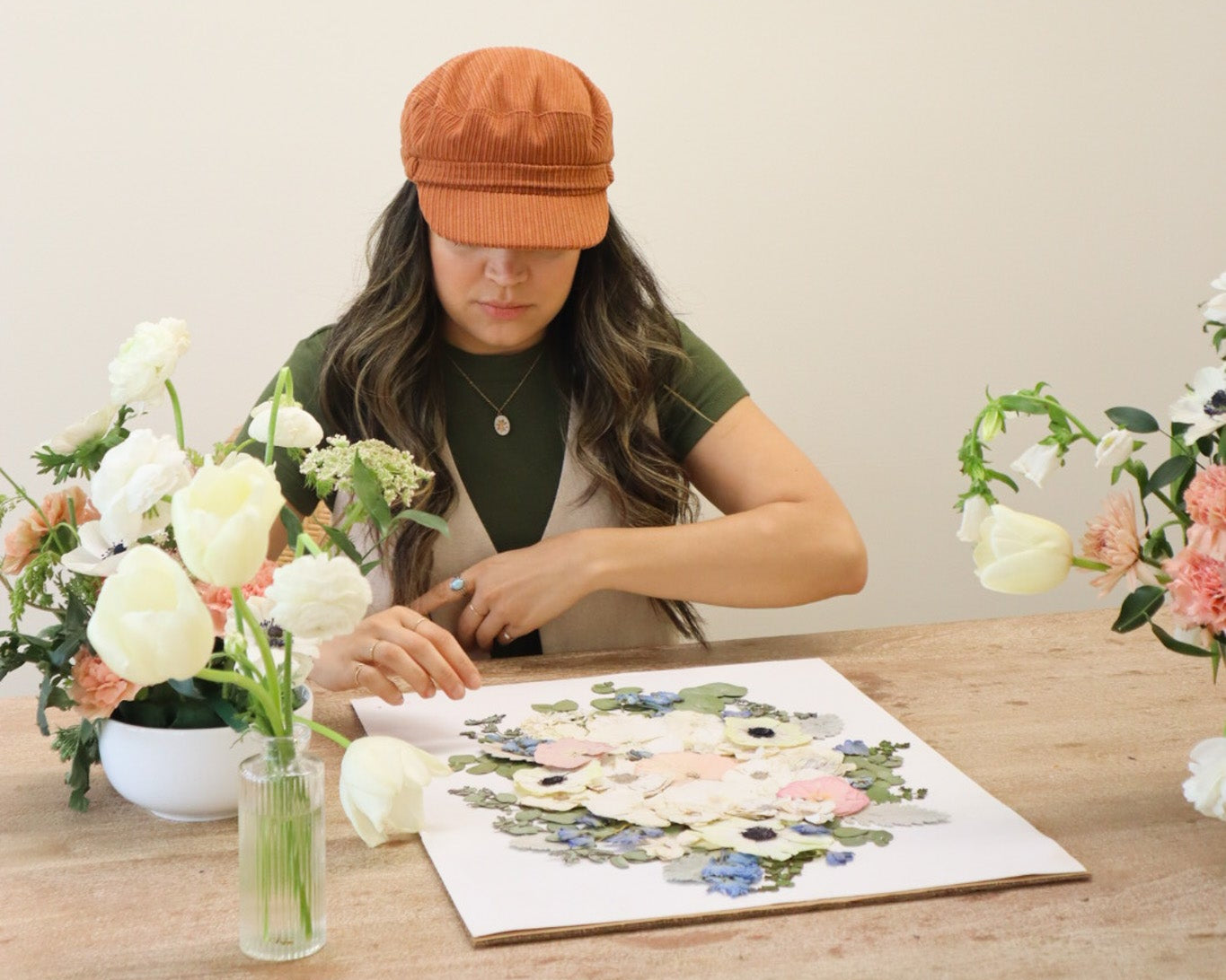 Person wearing an orange cap working on a floral design at a table with flowers and art supplies.