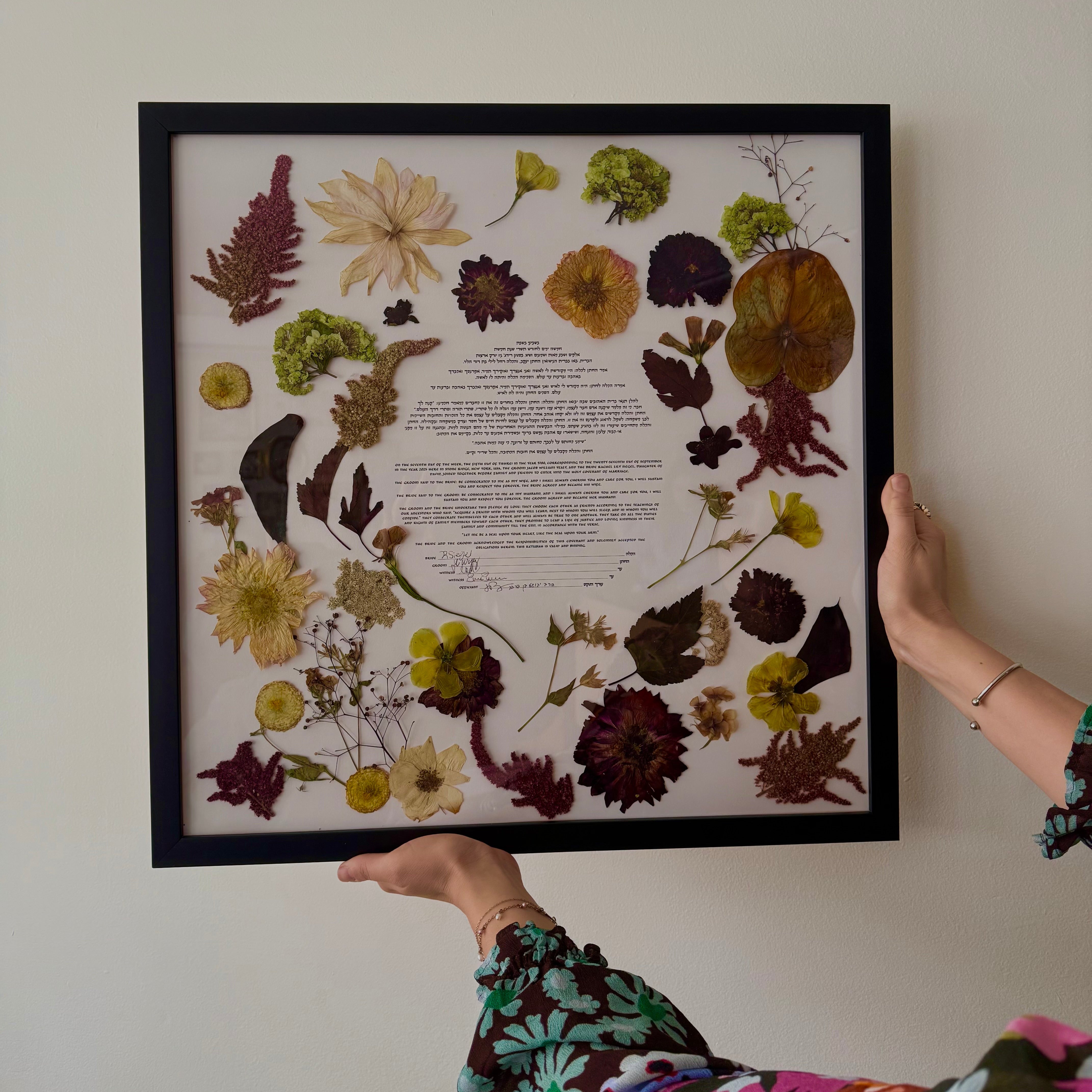 Framed ketubah with pressed flowers held by a person against a white wall