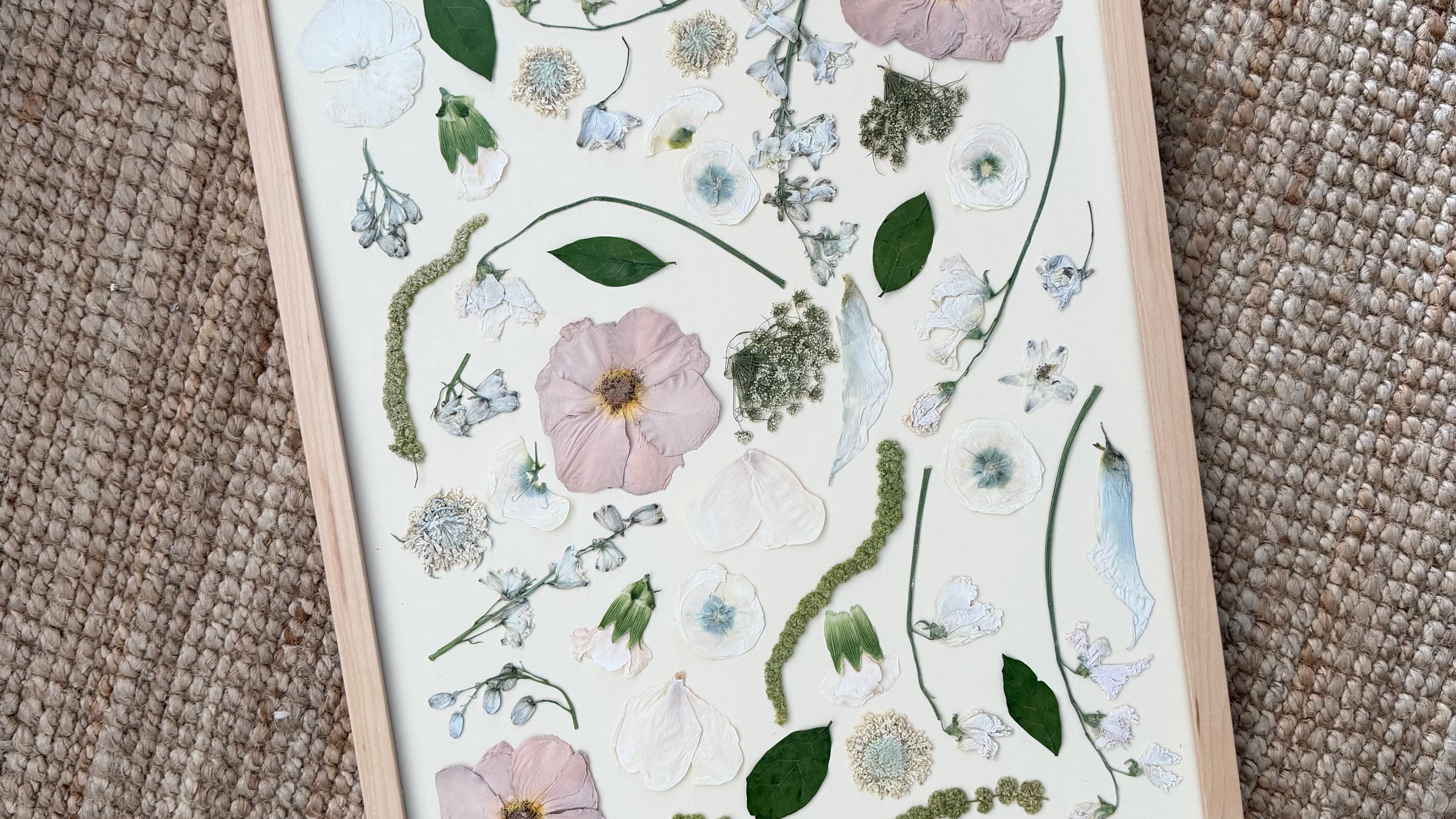 A framed botanical art print featuring pressed flowers and green leaves on a white background, set in a light wood frame and photographed on a textured beige woven rug.