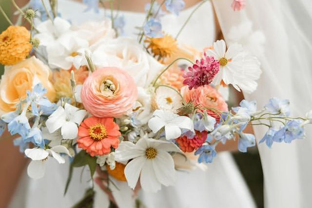 A close-up of a bride in a white wedding dress holding a vibrant, multicolored wildflower bouquet with peach, orange, blue, pink, and white flowers.