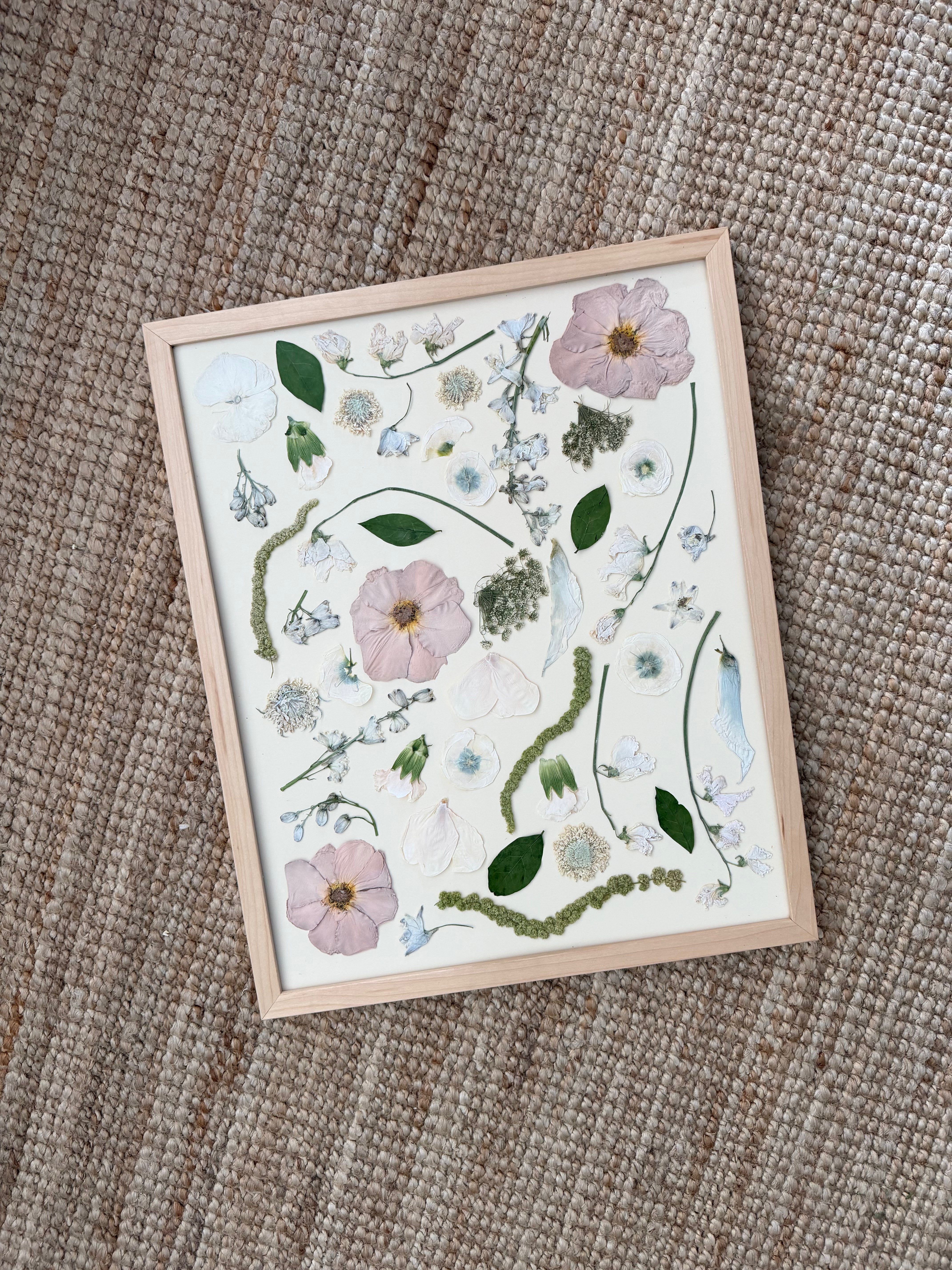 A framed botanical art print featuring pressed flowers and green leaves on a white background, set in a light wood frame and photographed on a textured beige woven rug.